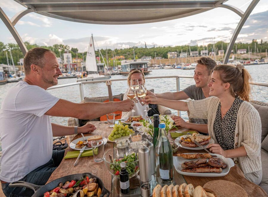 Gemeinsames Anstoßen mit Weingläsern an einem gedeckten Tisch auf einem Grillboot am Geiseltalsee