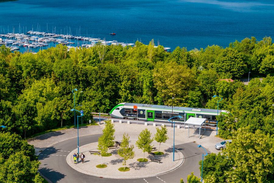Ein grüner Triebwagen der S-Bahn-Linie S11 steht am Bahnhof Mücheln am Geiseltalsee. Im Hintergrund sind der See mit zahlreichen Segel- und Hausbooten sowie der Yachthafen zu sehen, umgeben von viel Wald und Natur.