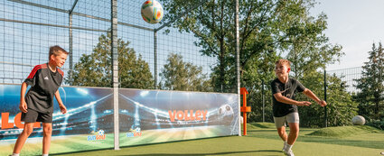 Zwei Jungen spielen auf einer Wiese Fußball. Der Ball befindet sich zentral in der Mitte und die Jungen stehen jeweils links und rechts und befinden sich in einer Schieß-Bewegung.