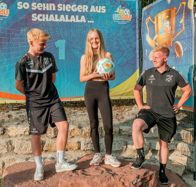 Ein Mädchen hält einen Fussball und freut sich, links und rechts stehen zwei Jungs, im Hintergrund steht auf der Plakatwand "So sehen Sieger aus"