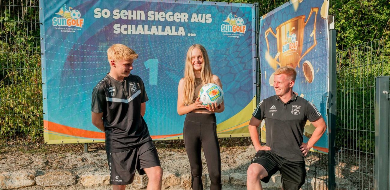 Ein Mädchen hält einen Fussball und freut sich, links und rechts stehen zwei Jungs, im Hintergrund steht auf der Plakatwand "So sehen Sieger aus"
