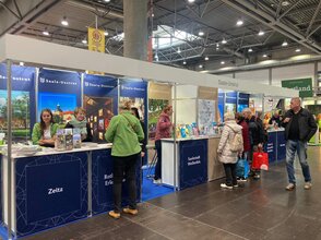 Es ist ein blau-goldener Messestand zusehen. Sowohl vor als auch hinter dem Messestand stehen Personen.