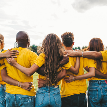 Gruppe junger Menschen in gelben T-Shirts steht im Freien mit verschränkten Armen um die Schultern und blickt gemeinsam in die Landschaft.