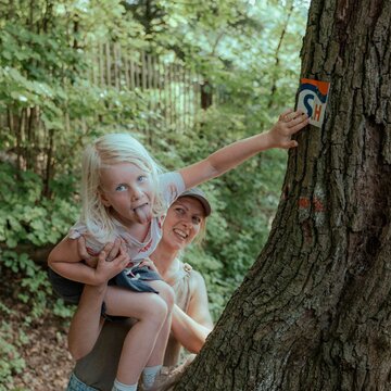 Frau hält ein blondes Mädchen fest, das zum Spaß die Zunge rausstreckt. Das Mädchen hält sich an einem dicken Baumstamm fest, genau dort, wo sich der Wegweiser des Wanderweges mit den Buchstaben SH befindet.