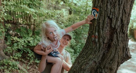 Frau hält ein blondes Mädchen fest, das zum Spaß die Zunge rausstreckt. Das Mädchen hält sich an einem dicken Baumstamm fest, genau dort, wo sich der Wegweiser des Wanderweges mit den Buchstaben SH befindet.