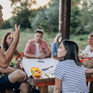 Gruppe junger Erwachsener sitzt im Freien an einem Tisch, während ein Mann etwas erklärt und die anderen aufmerksam zuhören.