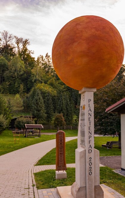 Wegweiser Tautenburger Planetenpfad: Eine rote Kugel thront auf einem schmalen, eckigen Pfeiler mit der Aufschrift Plantenpfad. Daneben ist ein Weg und im Hintergrund ein Wald zu sehen.