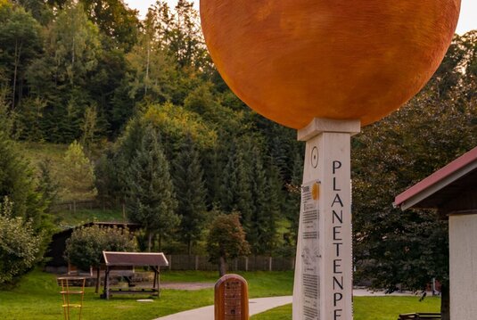 Wegweiser Tautenburger Planetenpfad: Eine rote Kugel thront auf einem schmalen, eckigen Pfeiler mit der Aufschrift Plantenpfad. Daneben ist ein Weg und im Hintergrund ein Wald zu sehen.