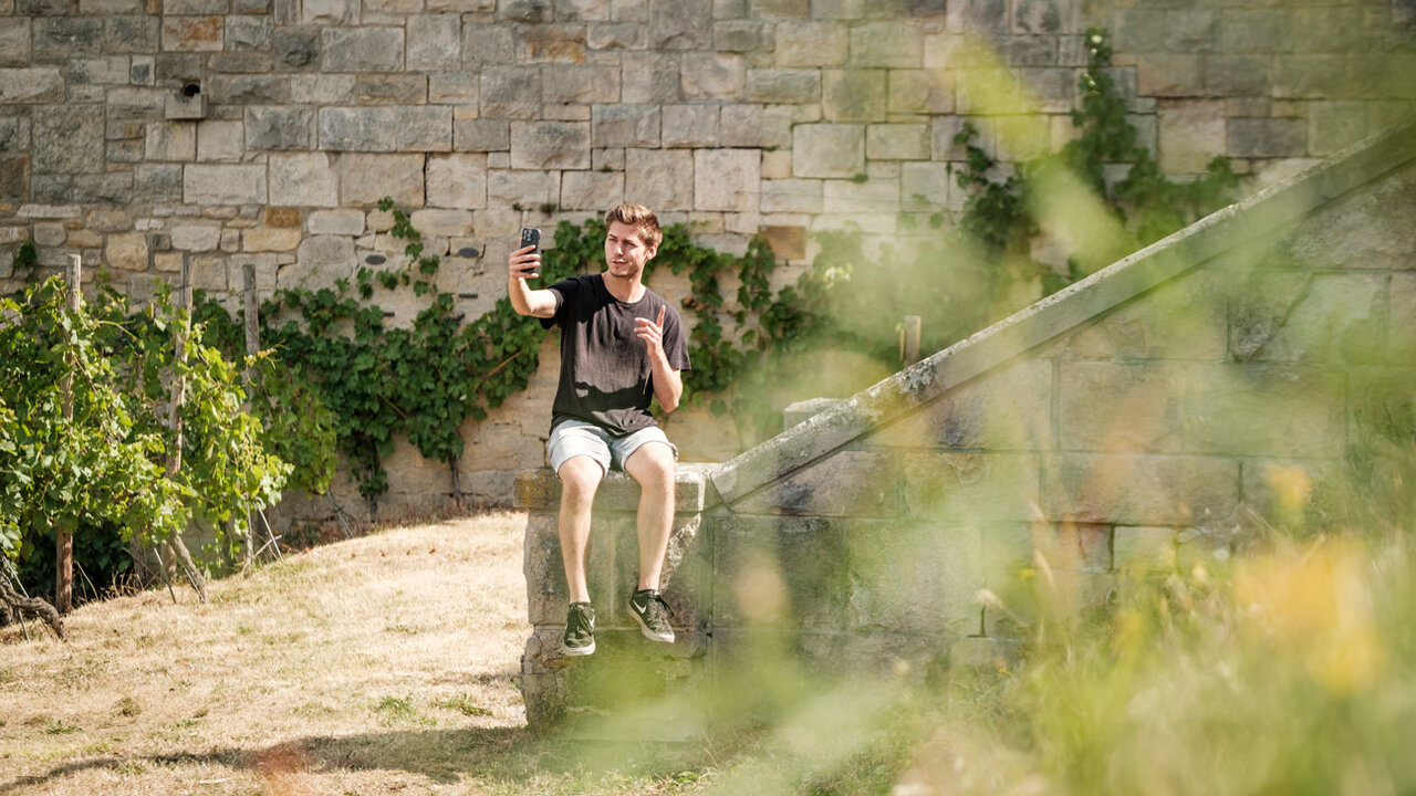 youtube Video Thumbnail: Junger Mann sitzt auf Mauer und filmt sich selbst im Garten vom Hotel Edelacker