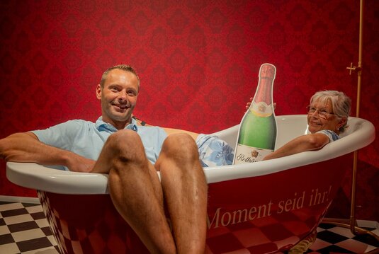 ein mann und eine Frau sitzen in einer Wanne, dahiner eine rote Wand, auf der Wanne steht "Der Moment seid ihr"