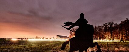 Stahlstele, das einen Reiter auf einem Pferd zeigt, am Schlachtfeldpfad Lützen, dahinter in indirekter Beleuchtung das Museumsgebäude Museum Lützen 1632
