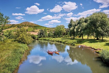 zwei Kanus auf einem Fluss, am Horizont ist ein Hügel mit Weinberg zu sehen, der Himmel ist blau mit wenigen Wolken, die sich im Wasser spiegeln, die Sonne scheint