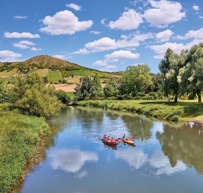 zwei Kanus auf einem Fluss, am Horizont ist ein Hügel mit Weinberg zu sehen, der Himmel ist blau mit wenigen Wolken, die sich im Wasser spiegeln, die Sonne scheint