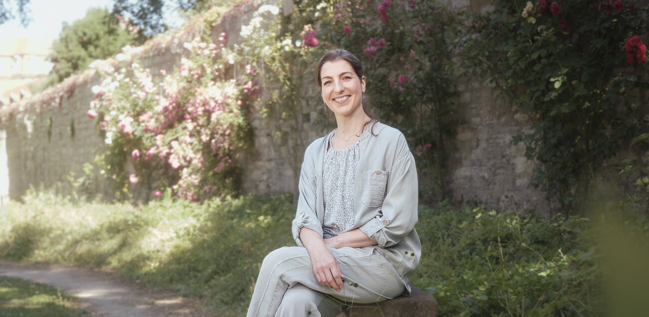 Nancy Weist sitzt strahlend im Garten mit blühenden Rosen im Hintergrund