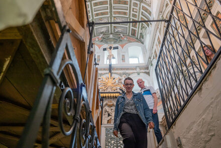 frau steht vor einem Treppenabgang, über ihr ist die reich verzierte Kassettendecke de rSchlosskirche Weißenfels zu sehen