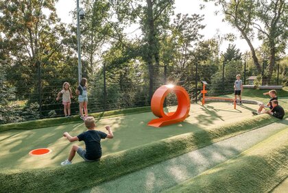 Ein Kind spielt Fußballgolf während andere hinter um die Station sitzen und dem Kind zuschauen.