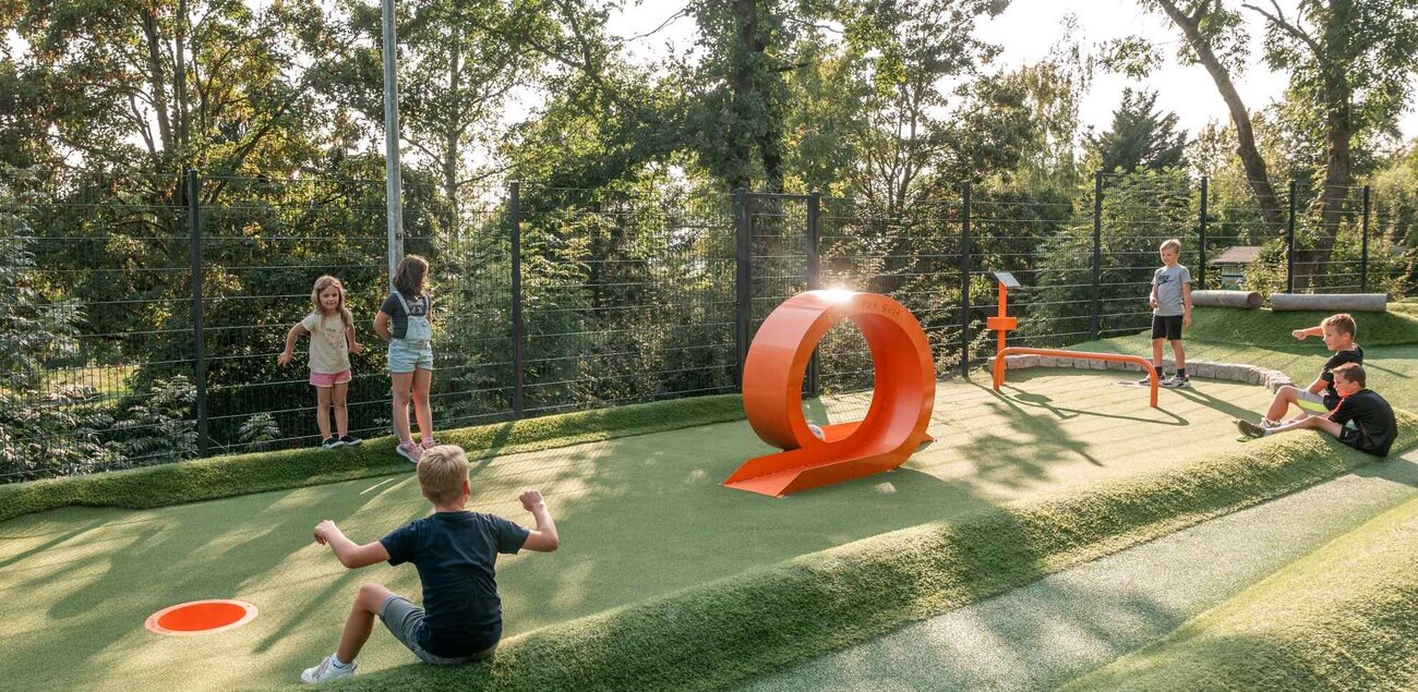 Ein Kind spielt Fußballgolf während andere hinter um die Station sitzen und dem Kind zuschauen.