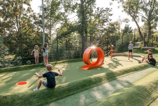 Ein Kind spielt Fußballgolf während andere hinter um die Station sitzen und dem Kind zuschauen.