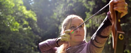 Eine Frau steht im Wald und hält einen Bogen mit einem Pfeil. Der Bogen ist bereits mit dem Pfeil gespannt.