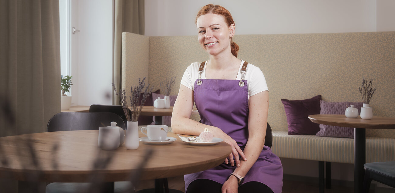 Annalena Jänicke sitzt lächelnd in ihrer violetten Schürze an einem Tisch in ihrem Café Lavendelblüte