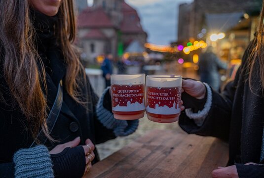 zwei Frauen stoßen mit Glühweintassen an, auf denen der Schriftzug Querfurter Weihnachtszauber sowie Schneeflocken aufgedruckt sind