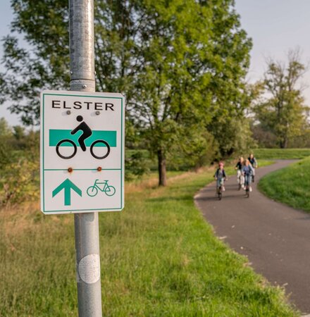Radwegeschild zum elsterradweg, im Hintergrund fährt eine Familie auf dem Radweg entlang