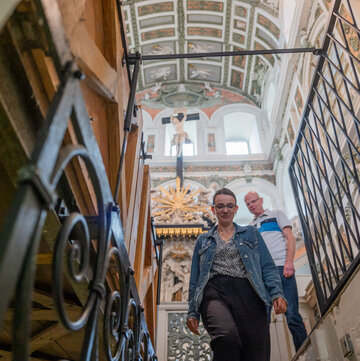 frau steht vor einem Treppenabgang, über ihr ist die reich verzierte Kassettendecke de rSchlosskirche Weißenfels zu sehen