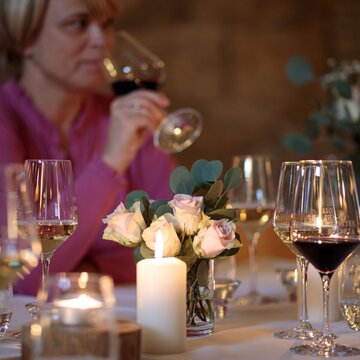 festlich gedeckter Tisch mit Blumenbouquet, leuchtender Kerze, Wein- und Wassergläsern, im Hintergrund nippt eine Frau an einem Gals Wein