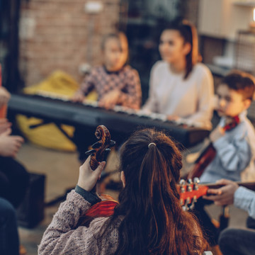 Kinder und Erwachsene sitzen in einem Raum zusammen und musizieren mit Geige, Gitarre und Keyboard im Gruppenunterricht.
