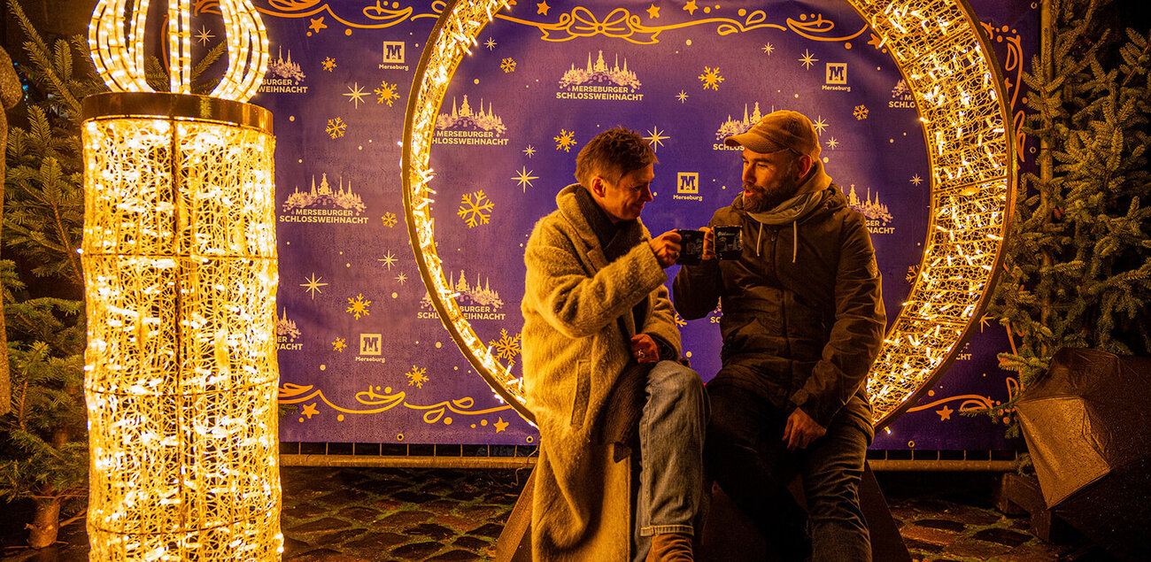 Ein mann und eine Frau stoßen mit Glühwein an. Sie sitzen in einem mit Lichterketten beleuchtetem Rondell, daneben eine mit Lichterketten beleuchtete, menschengroße Kerze