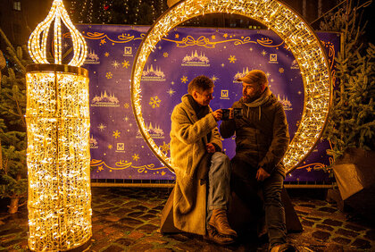Ein mann und eine Frau stoßen mit Glühwein an. Sie sitzen in einem mit Lichterketten beleuchtetem Rondell, daneben eine mit Lichterketten beleuchtete, menschengroße Kerze