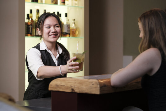Portrait von Azubi Sydney Gunawan hinter der Bar beim Servieren eines Cocktails