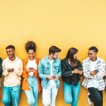 Sechs Jugendliche lehnen an einer gelben Wand und schauen lachend auf ihre Smartphones.