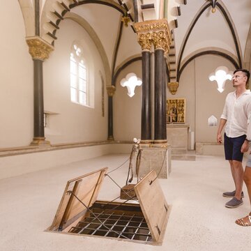 Ein Paar besucht die Doppelkapelle Schloss Neuenburg. Es ist ein weißer Raum mit romanischen Fenstern und einer geöffneten Bodenklappe.