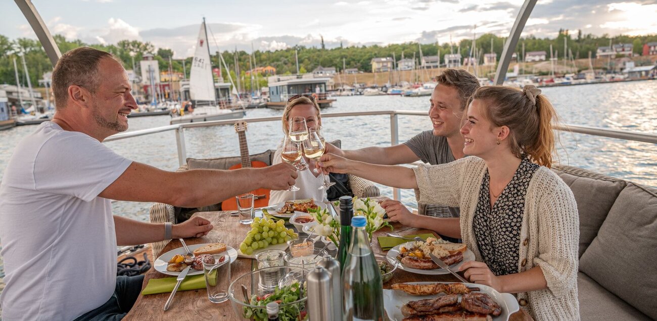 Vier Personen sitzen auf einem Boot und auf dem Tisch ist Essen zu sehen. Die Personen stoßen gemeinsam mit Weingläsern an.