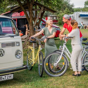 Zwei Frauen und ein Mann stehen mit Fahrrädern in eine rGruppe zusammen und unterhalten sich. Um sie herum sind mehrere Wohnmobile zu sehen und ein überdachter Picknickplatz zu sehen.