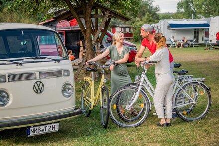 Zwei Frauen und ein Mann stehen mit Fahrrädern in eine rGruppe zusammen und unterhalten sich. Um sie herum sind mehrere Wohnmobile zu sehen und ein überdachter Picknickplatz zu sehen.