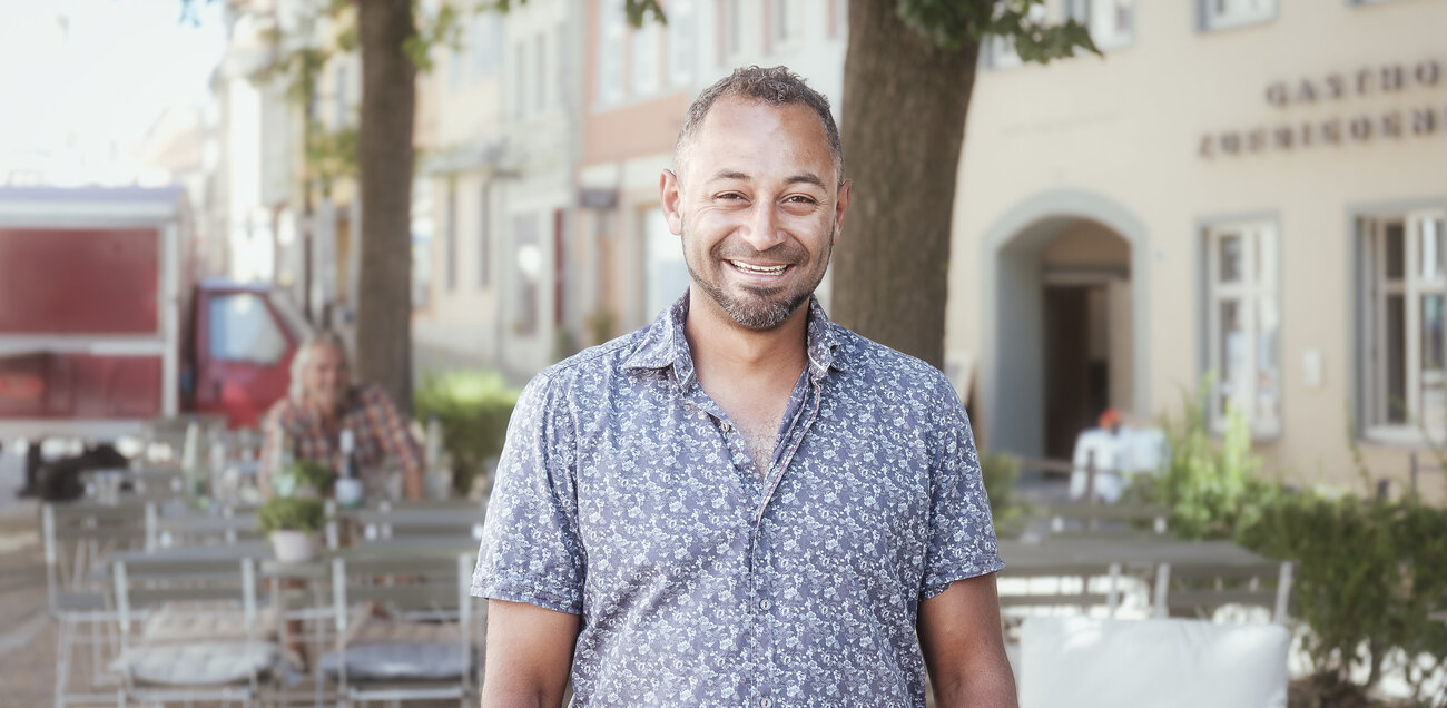 Manuel Flechtner steht vor einer Weinbar unter einem Baum und strahlt in die Kamera