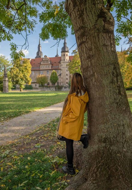 Frau lehnt im herbstlichen Schlosspark Merseburg am Baum und schaut verträumt Richtung Dom-Schloss-Ensemble