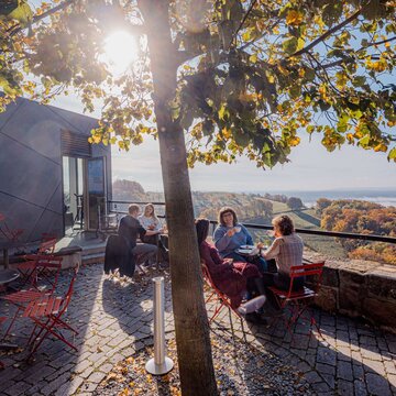 Personen sitzen an verschiedenen Tischen und trinken und essen dabei etwas. Im Hintergrund ist der Blick von der Leuchtenburg zu sehen.