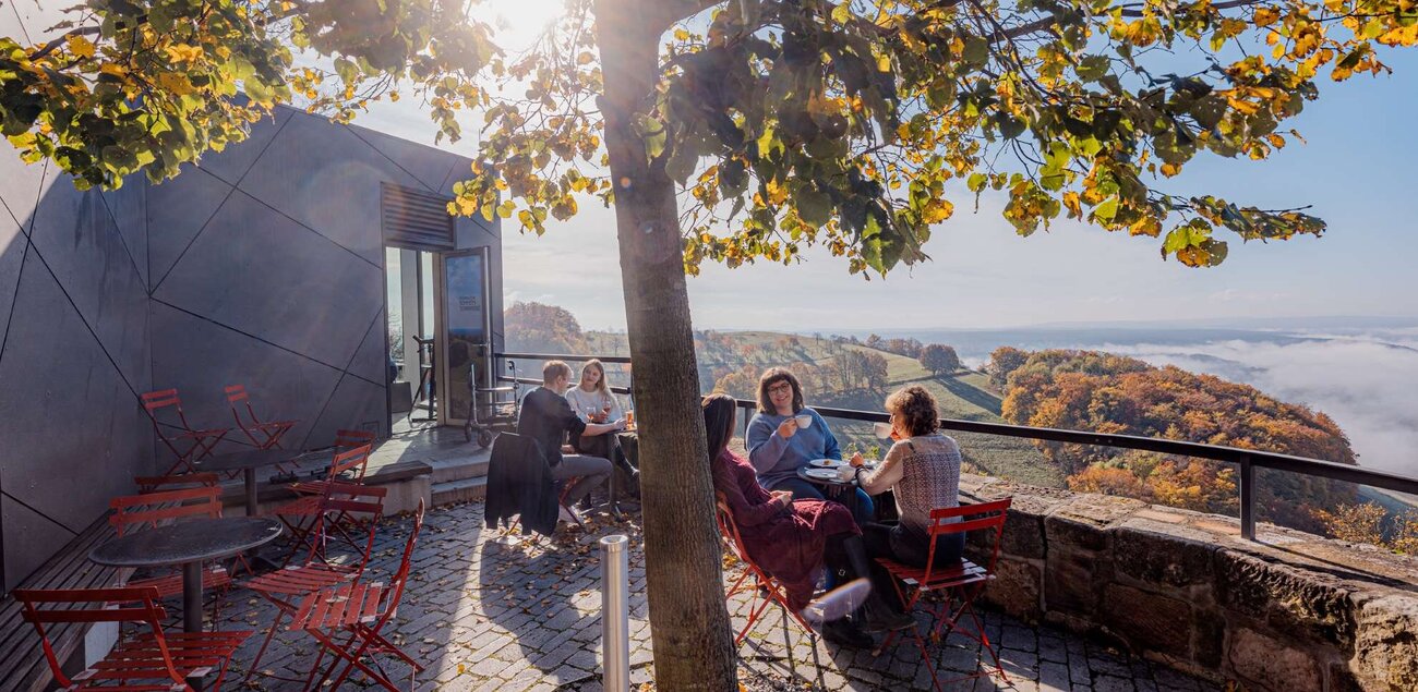 Personen sitzen an verschiedenen Tischen und trinken und essen dabei etwas. Im Hintergrund ist der Blick von der Leuchtenburg zu sehen.