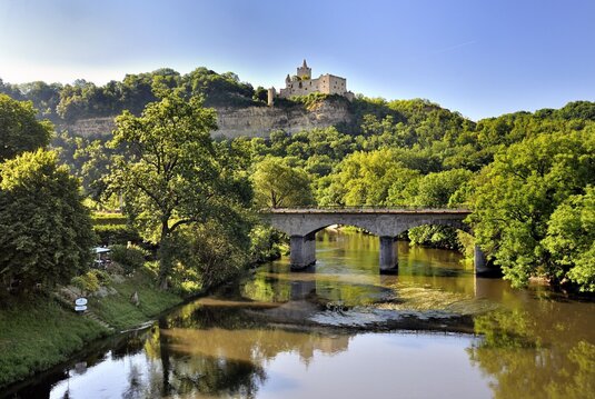 Eine Steinbrücke über dem Fluss Saale mit der Rudelsburg auf einem bewaldeten Hügel im Hintergrund.