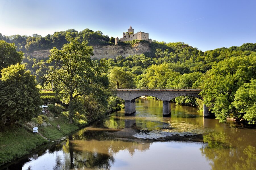 Eine Steinbrücke über dem Fluss Saale mit der Rudelsburg auf einem bewaldeten Hügel im Hintergrund.