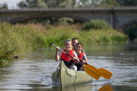 Zwei Frauen und ein Mann sitzen in einem Kanu, das sich mitten auf einem Fluss befindet. Jeder hat ein Paddel und taucht es ins Wasser.