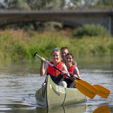 Zwei Frauen und ein Mann sitzen in einem Kanu, das sich mitten auf einem Fluss befindet. Jeder hat ein Paddel und taucht es ins Wasser.