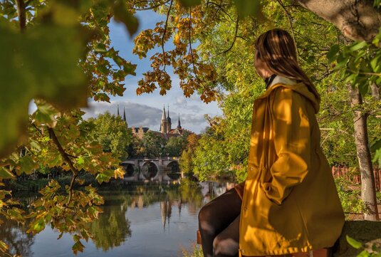 Frau in gelbem Herbstmantel sitzt an der Saale und schaut auf die Türme von Dom und Schloss Merseburg. Vor ihr fließt der Fluss, um sie herum hängen viele Zweige mit buntem Laub.