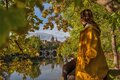 Frau in gelbem Herbstmantel sitzt an der Saale und schaut auf die Türme von Dom und Schloss Merseburg. Vor ihr fließt der Fluss, um sie herum hängen viele Zweige mit buntem Laub.