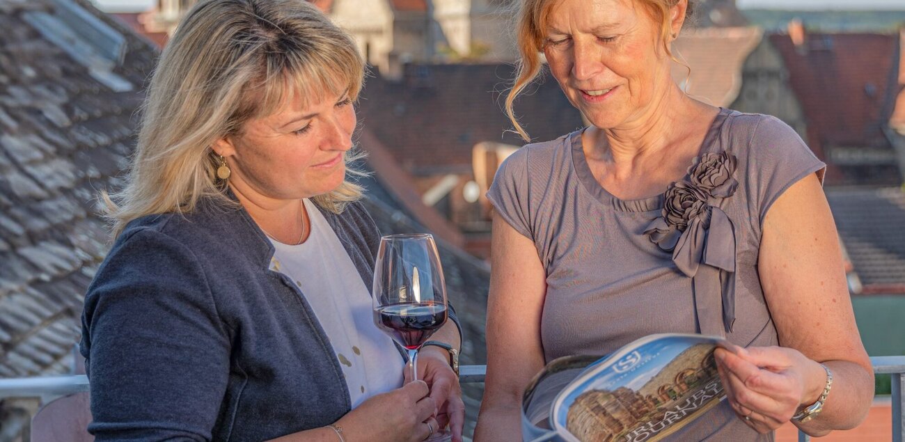 Zwei Frauen schauen sich ein Prospekt an. Eine der Frauen hält dabei noch ein Weinglas. Im Hintergrund sind Teile des Naumburger Doms zusehen.