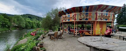 Zu sehen ist die "Wünsch dir was"-Bar, welche ein buntes Karussell ist. Um die Bar sitzen mehrere Menschen. Links auf dem Bild ist ein Fluss zu sehen, wo gerade Leute mit dem Kanu ankommen.