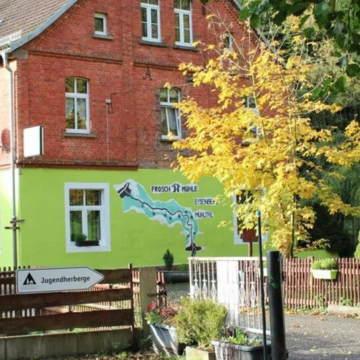 Jugendherberge in einem rot-grünen Backsteingebäude am Waldrand, mit Gartenzaun, Wegweiser und herbstlichen Bäumen im Vordergrund.